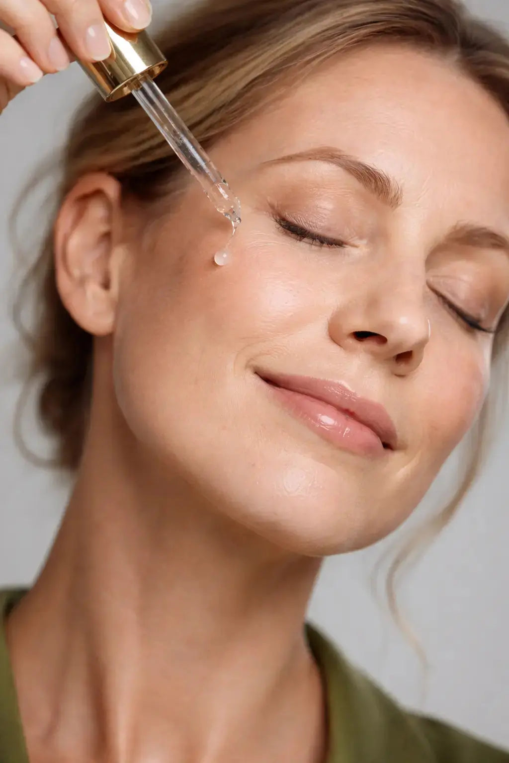Lady smiling applying face serum to her face. She is wearing a green top has highlighted blond hair