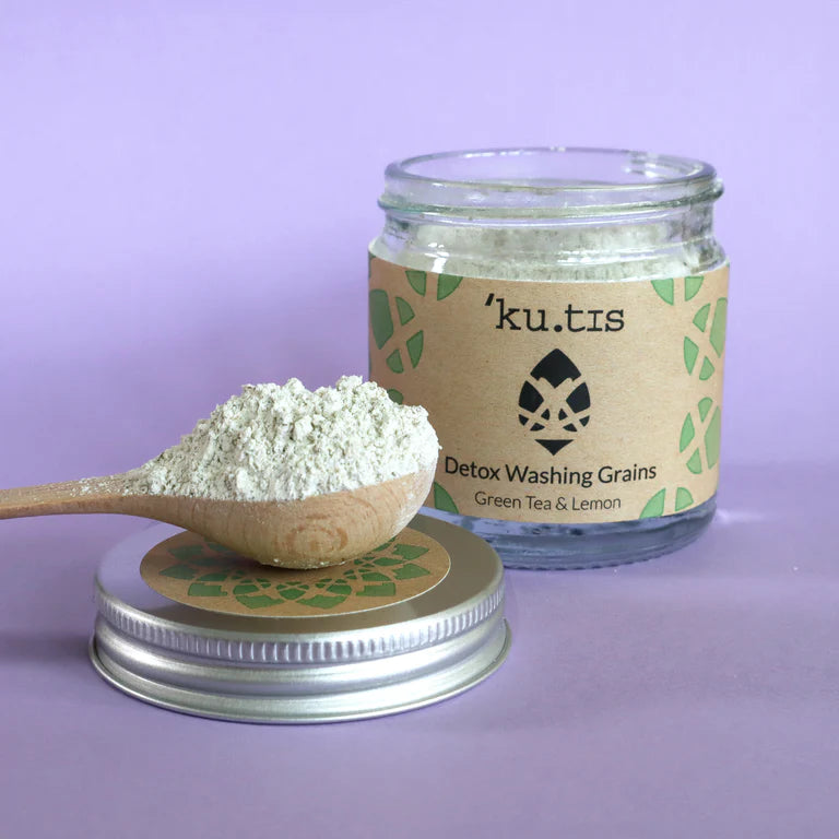 Jar of 'ku.tis' Detox Washing Grains with a wooden spoon filled with powder on a purple background, lid off., front