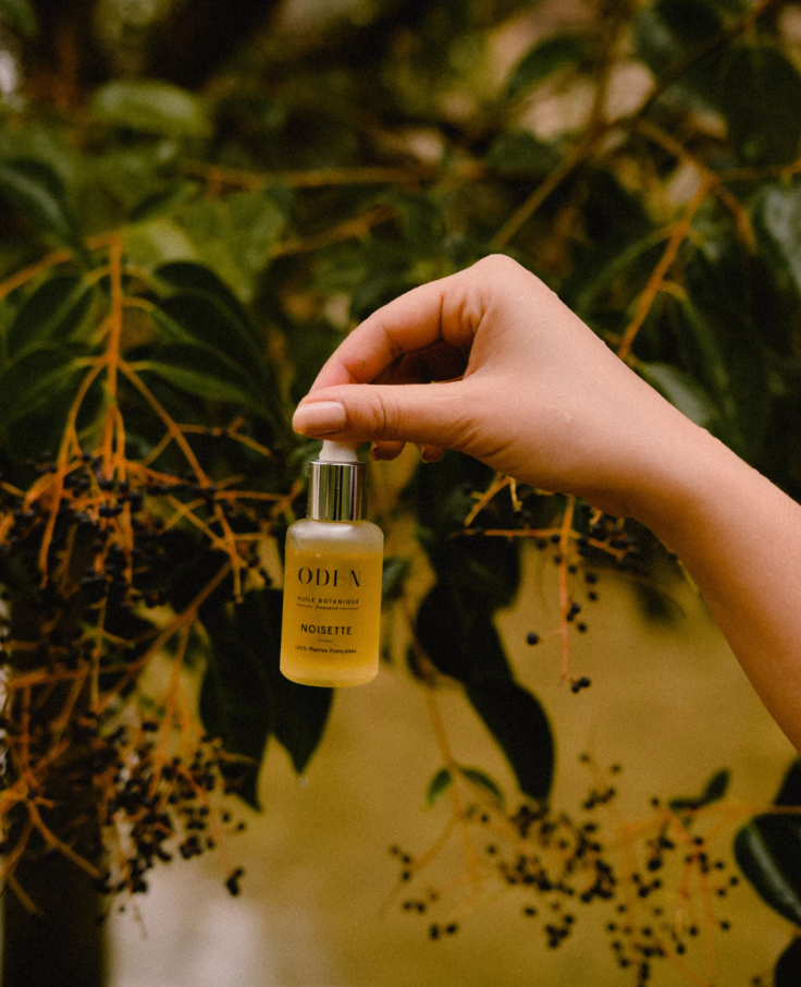 Hand holding a small bottle labelled 'ODEN' against a natural background with green leaves.