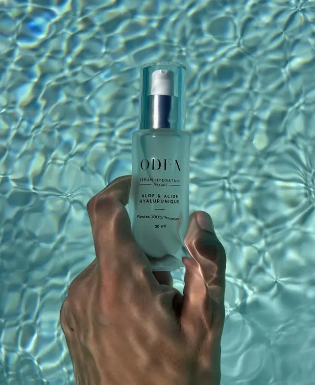 Hand holding a bottle of ODEN skincare product against a water background