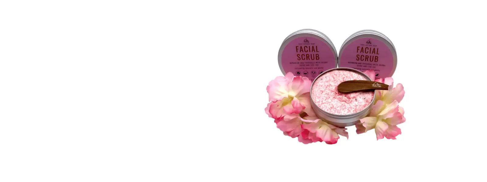 Cosy cottage facial scrub in pink tin with pink flowers around it. On a white background.