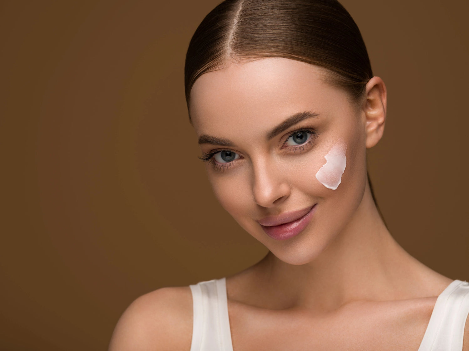 lady smiling at the camera with a patch of face cream on her cheek. Brown background