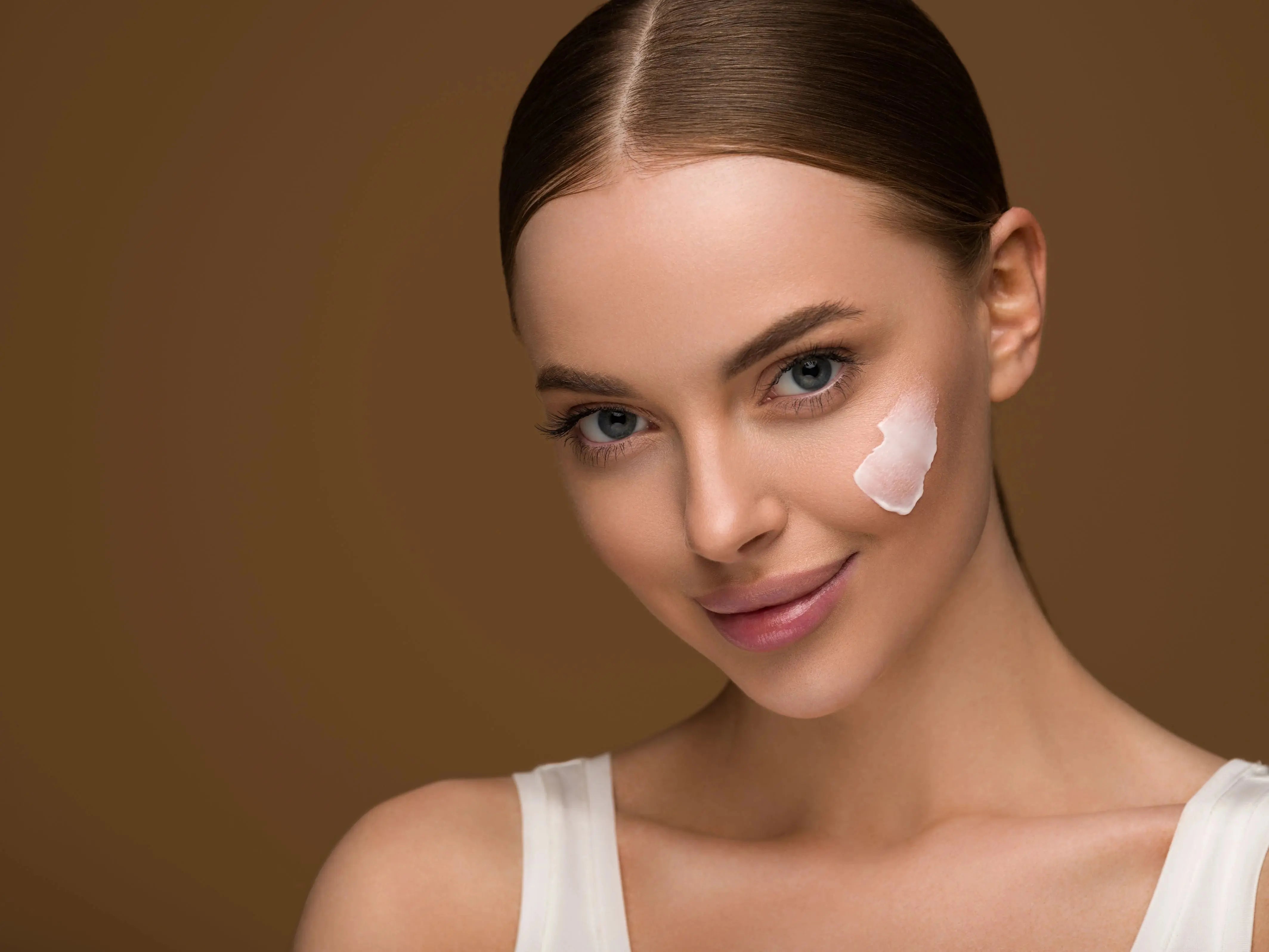 lady smiling at the camera with a patch of face cream on her cheek. Brown background
