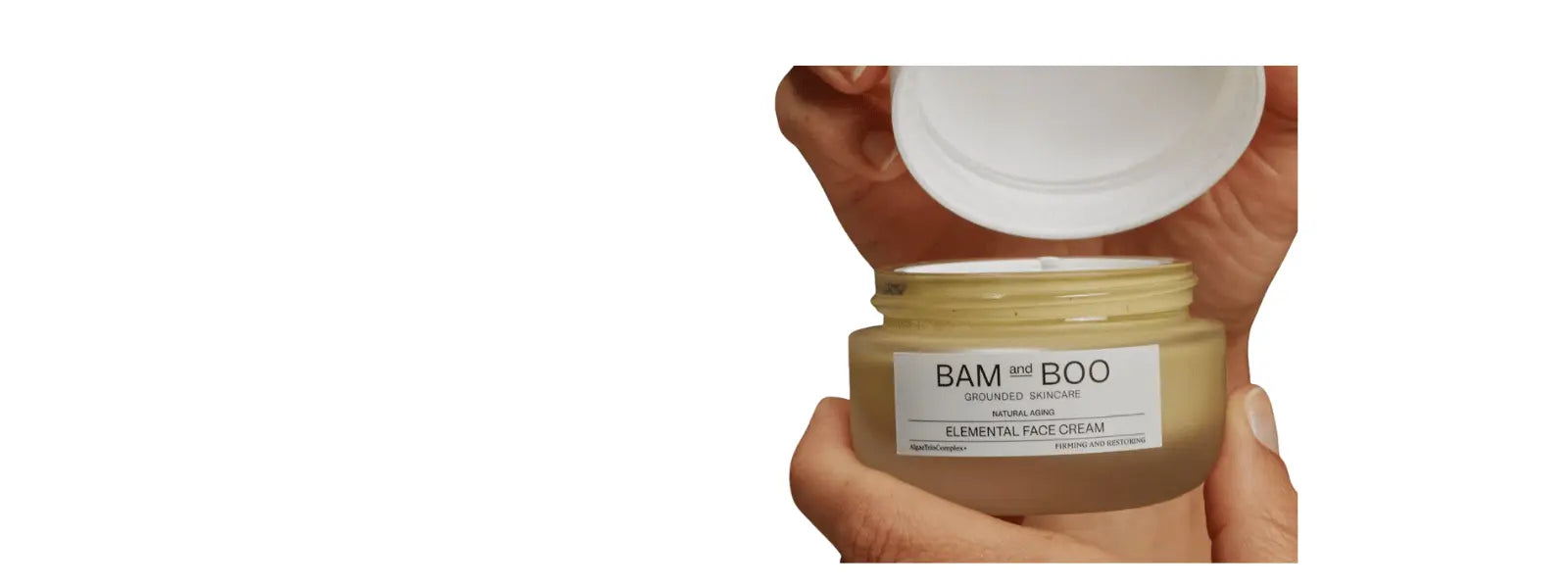 Hand holding Bam and Boo anti-ageing face cream, jar open, white lid on white bacground.