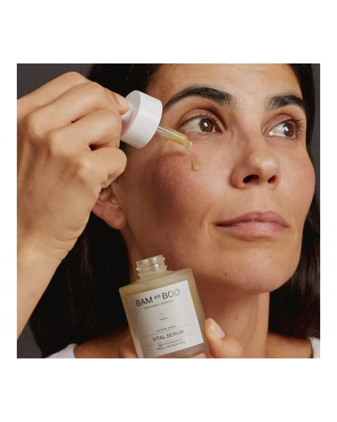 Woman applying a dropper of skincare product to her face with 'Bamford' bottle in hand.