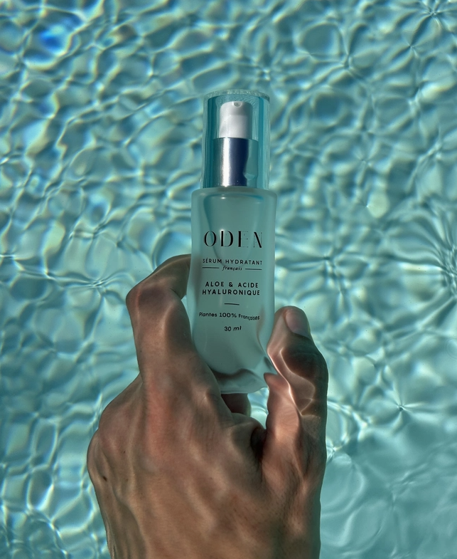 Hand holding a bottle of ODEN skincare product against a water background